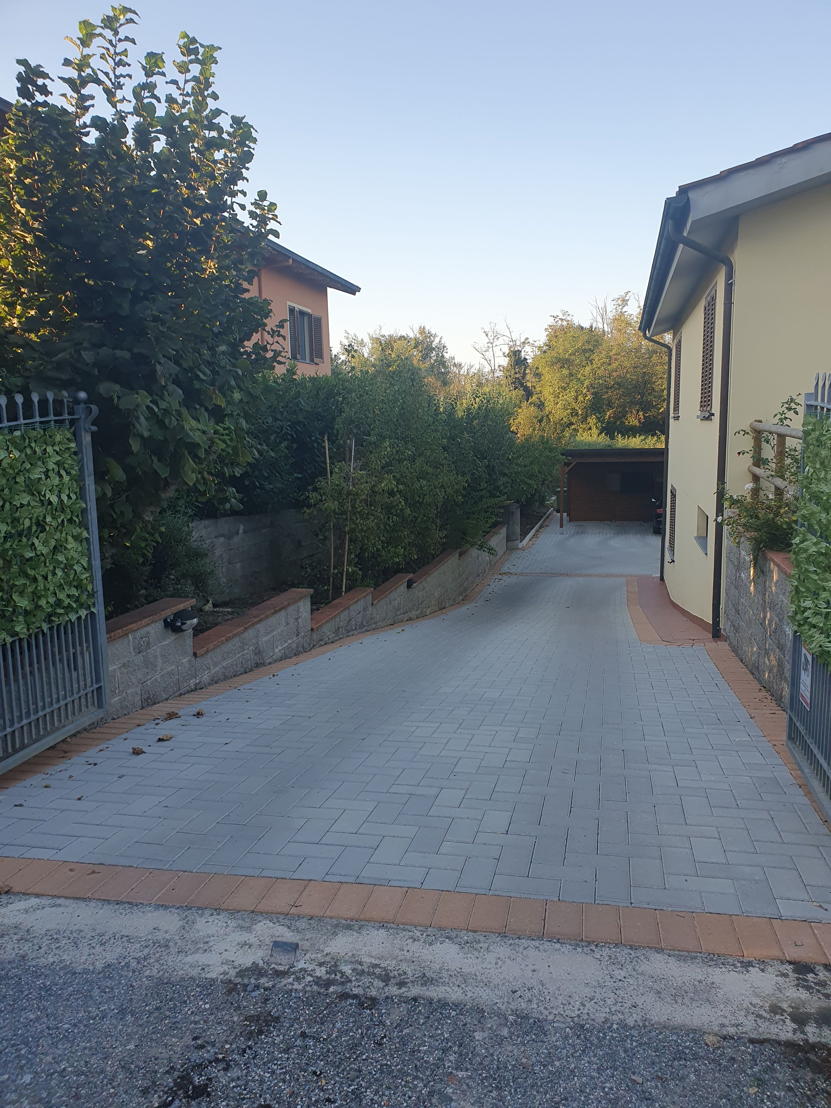 Ein gepflasterter Weg führt zu einem Wohnhaus mit Garage, umgeben von Grünflächen und einem Metallzaun.