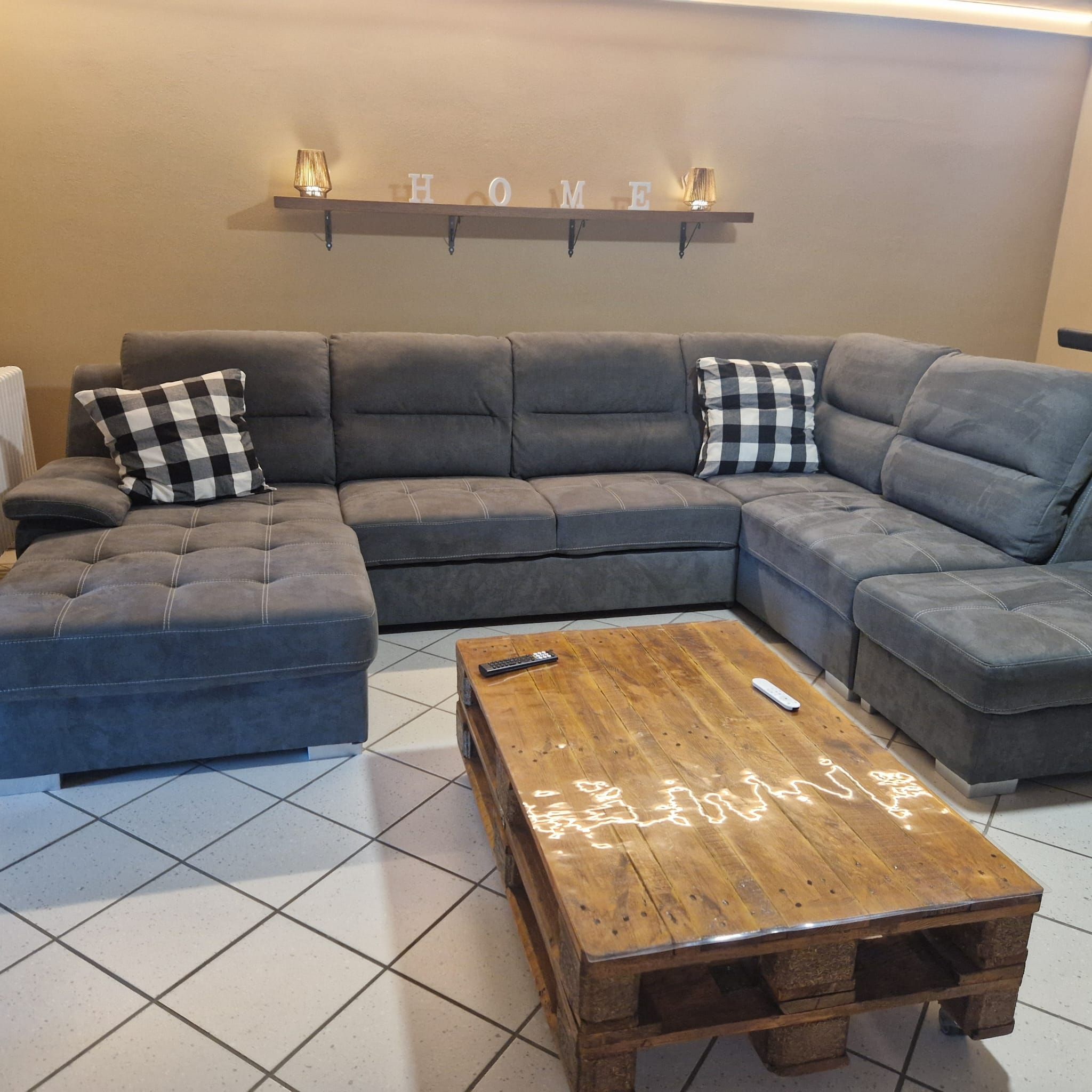 Um sofá modular com almofadas xadrez, uma mesa de centro de madeira e uma prateleira fixada na parede com a palavra 'HOME' em destaque.