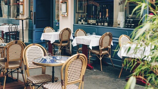 Een terras met rieten stoelen en tafels, een wijnrek op de achtergrond en een blauwe deur.