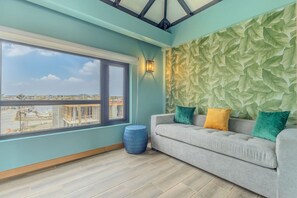 A room with a large window, a grey sofa with colorful cushions, and a textured wall. - LUX 1BR House Pool Gym 7-Min Walk Palm Beach - Porta Al Sole 8 by Bocobay (Noord)