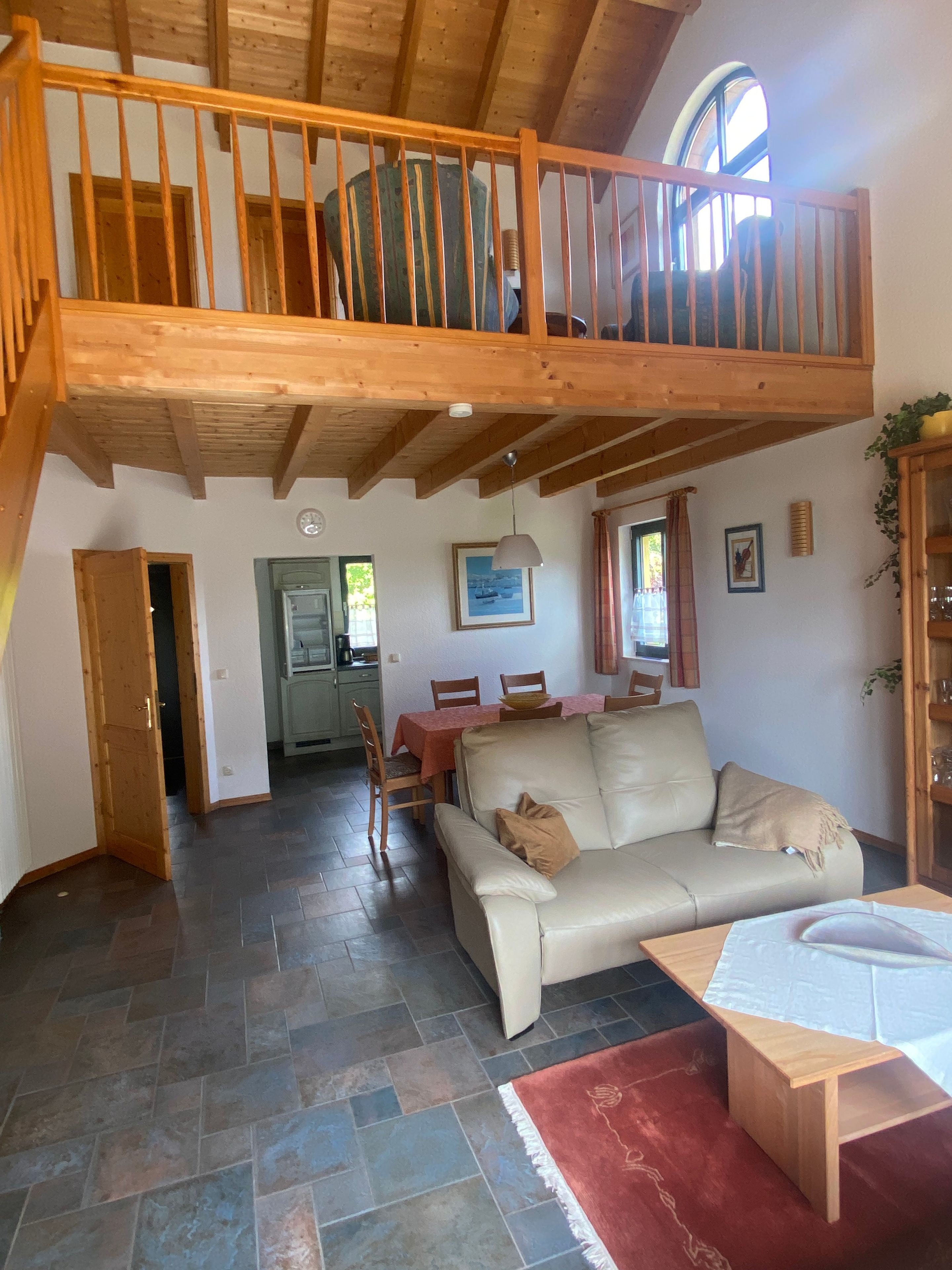 A spacious living area with a high wooden ceiling, a dining table, a sofa, and a staircase leading to an upper level.