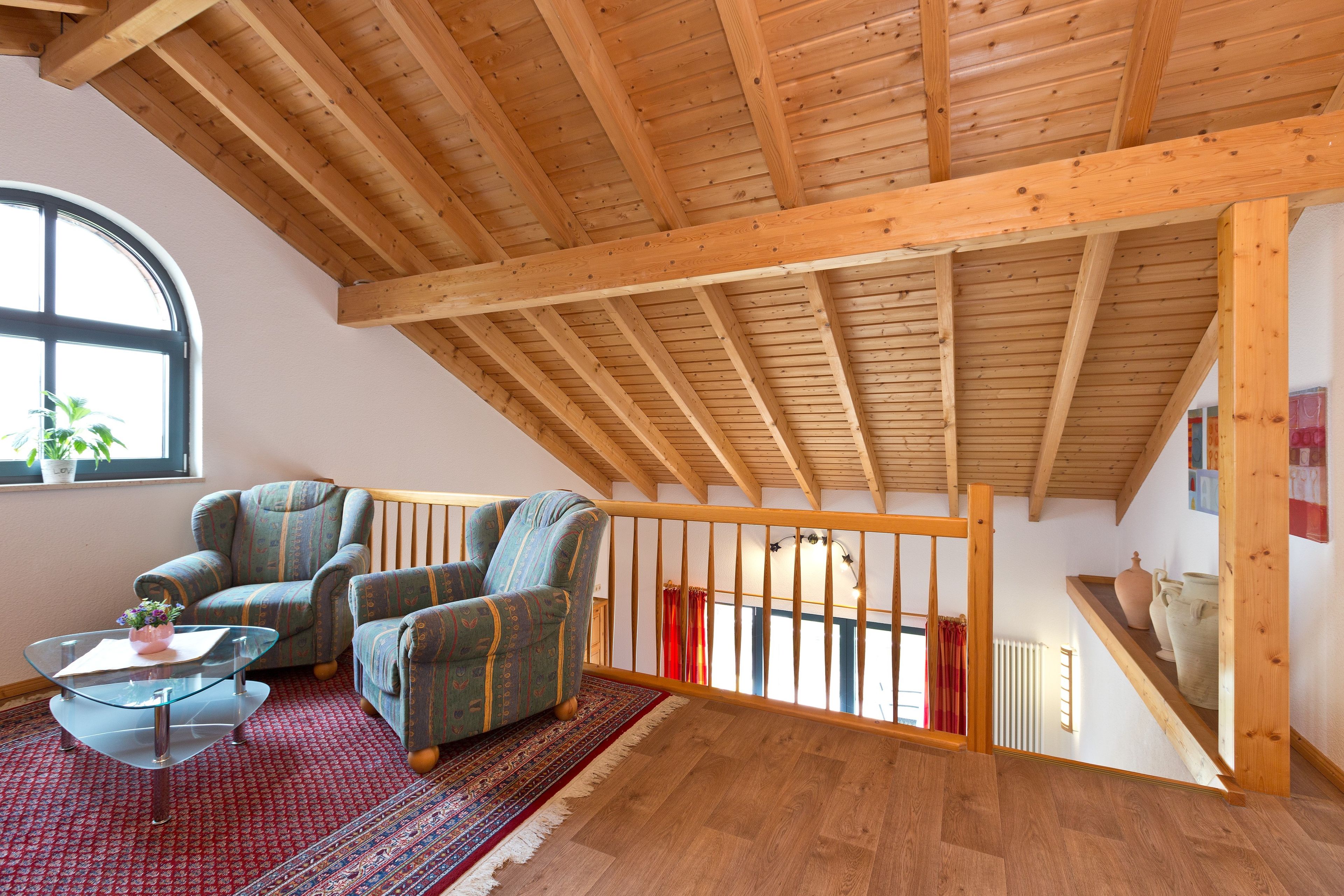 A spacious loft with exposed wooden beams, a large window, and a cozy seating area with patterned armchairs and a glass coffee table.