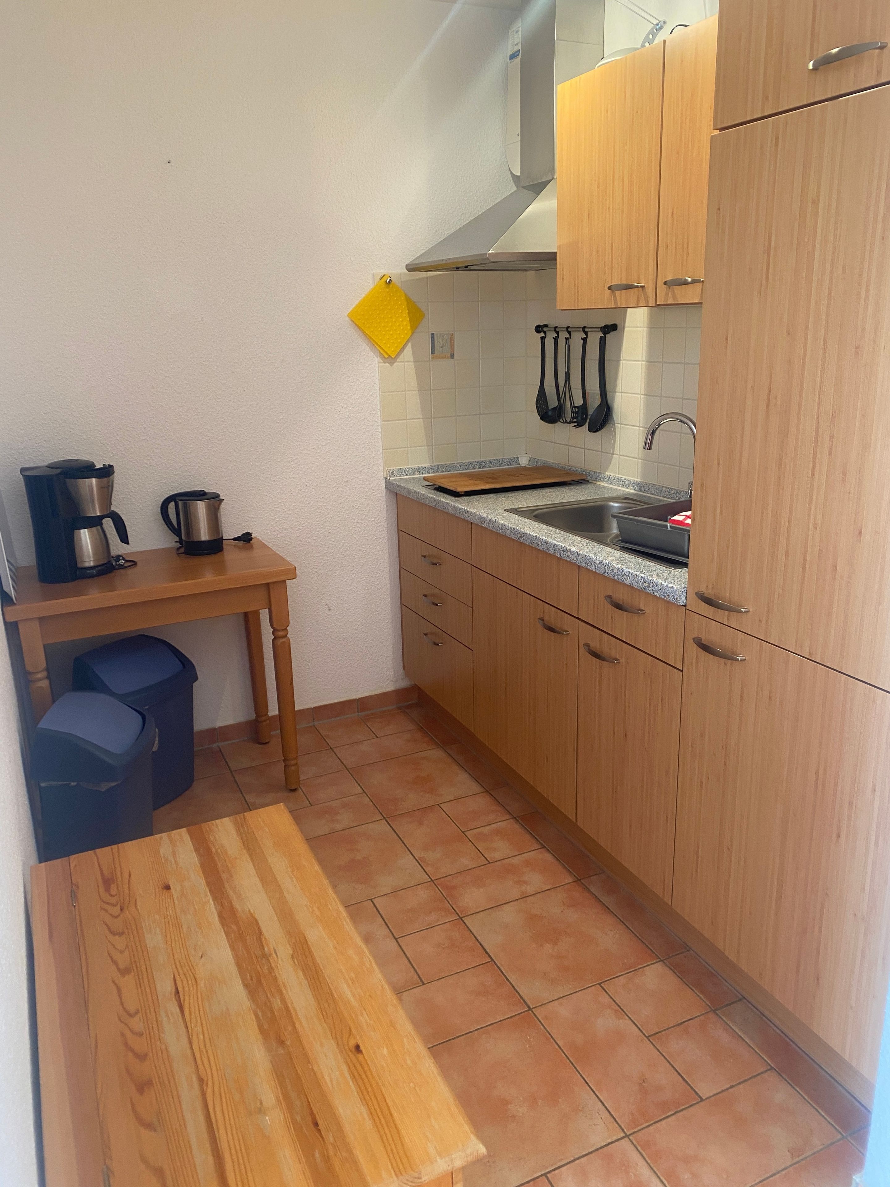 A compact kitchen with a sink, wooden cabinets, a countertop, and a small table with a coffee maker.