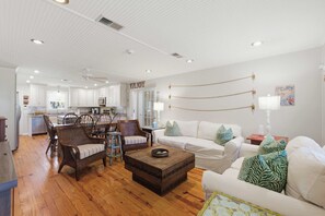 A spacious living room with a white sofa, a wooden coffee table, and wicker chairs. A kitchen is visible in the background with white cabinets and a dining area. - Edisto Escape (Edisto Island)