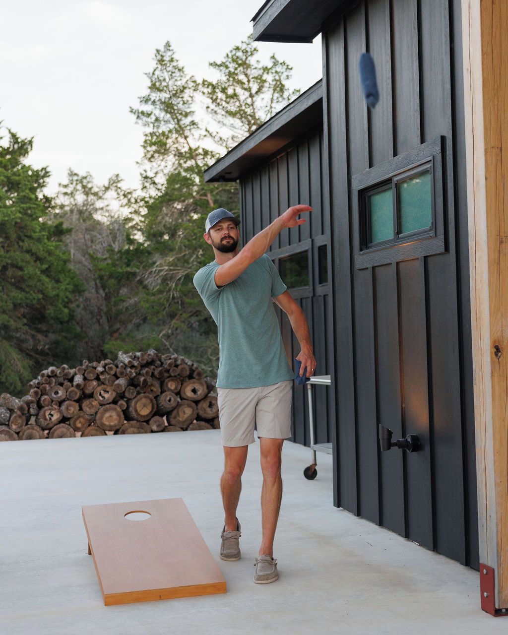 A person standing next to a building with a door, a pile of logs, and a wooden board on the ground.