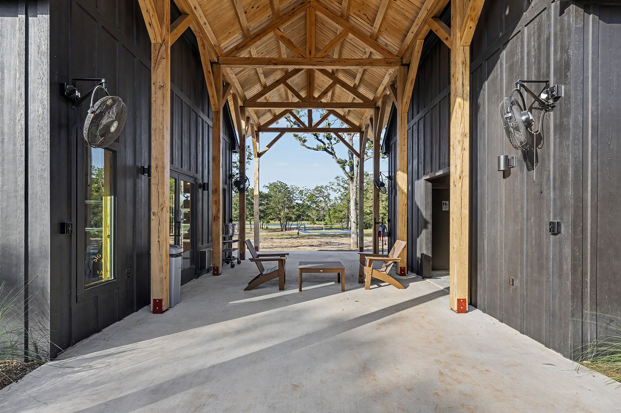 A covered outdoor area with a concrete floor, wooden benches, and a view of a park with trees and a clear sky.