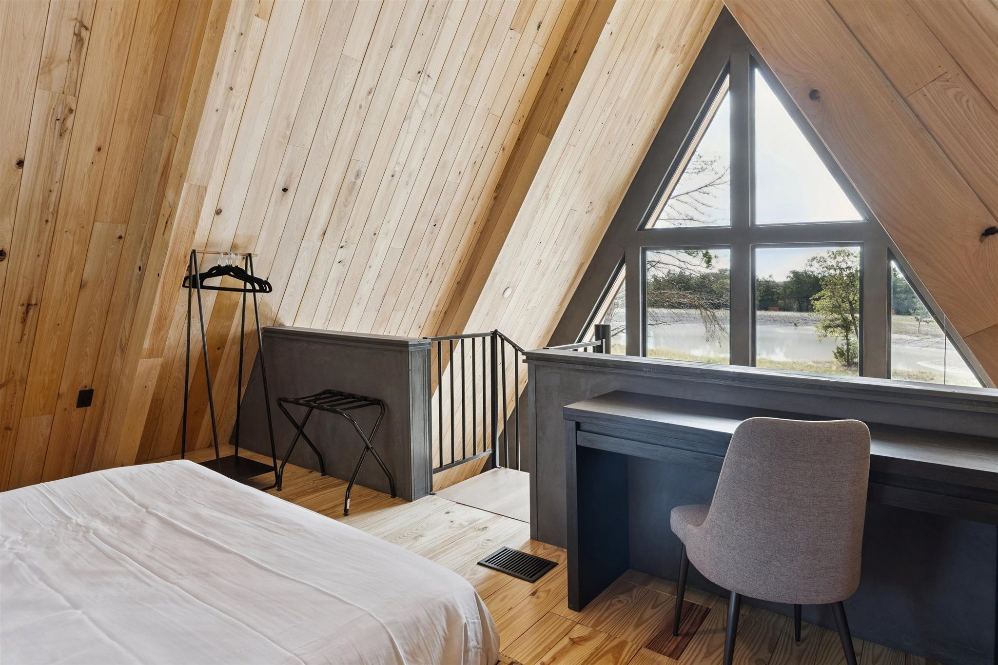 Loft bedroom with a queen‑size bed, floor‑to‑ceiling windows that frame sweeping lake views through the A‑frame cabin, and a sleek desk perfect for work or journaling.