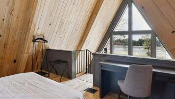 Loft bedroom with a queen‑size bed, floor‑to‑ceiling windows that frame sweeping lake views through the A‑frame cabin, and a sleek desk perfect for work or journaling.