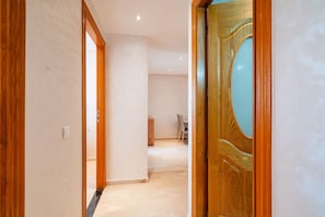 A hallway with a wooden door, a light-colored wall, and a visible dining area in the background. - Modern Escape in Gueliz |Terrasse| View | Parking (Marrakesh)