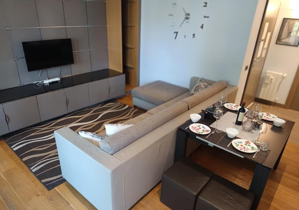 Un salon moderne avec un canapé d’angle, un téléviseur à écran plat fixé au mur, une table à manger dressée pour deux personnes avec des assiettes et des verres, et un tapis à motifs zébrés.
