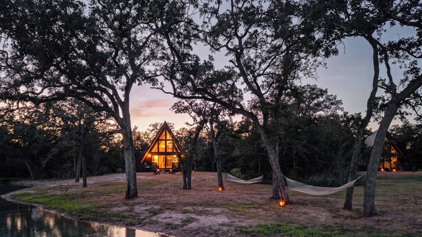 A cabin with a triangular roof surrounded by trees, a hammock, and a small pond. - A quiet A-frame escape in the Texas countryside, made for slowing down. (Ledbetter)
