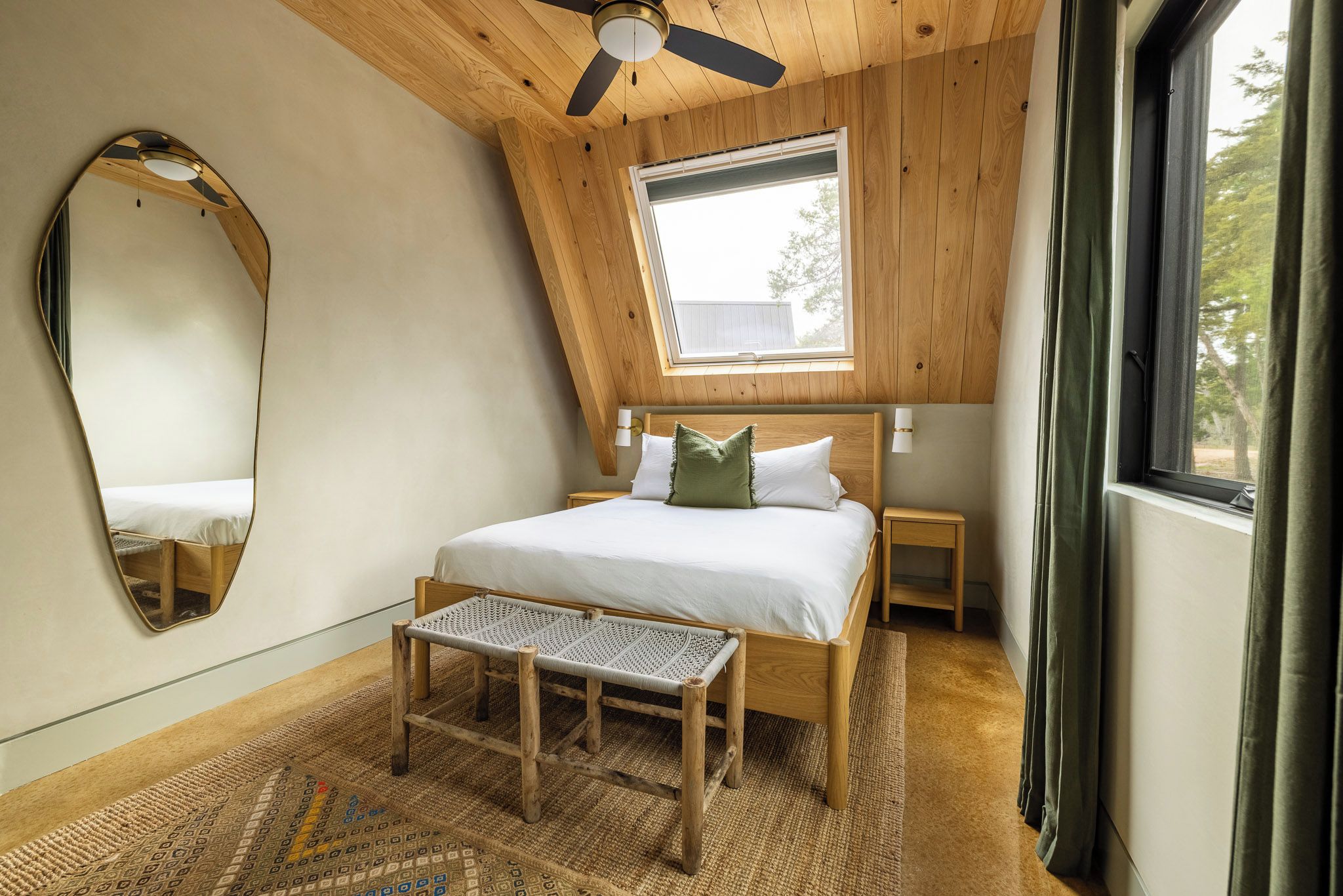 A bedroom with a sloped ceiling, a bed with pillows, a bench, a mirror, and a window.