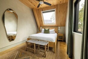 A bedroom with a sloped ceiling, a bed with pillows, a bench, a mirror, and a window.