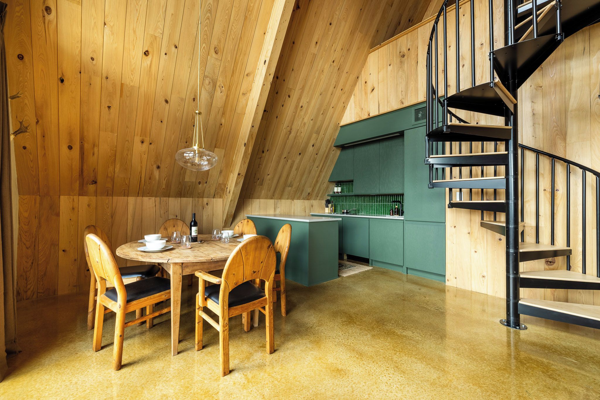 Uma área de refeições com mesa e cadeiras de madeira, uma cozinha com armários verdes e uma escada espiral preta.