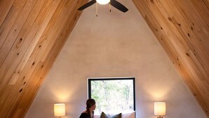 A bedroom with a sloped ceiling, a bed with a green blanket, two bedside lamps, and a ceiling fan. - A quiet A-frame escape in the Texas countryside, made for slowing down. (Ledbetter)