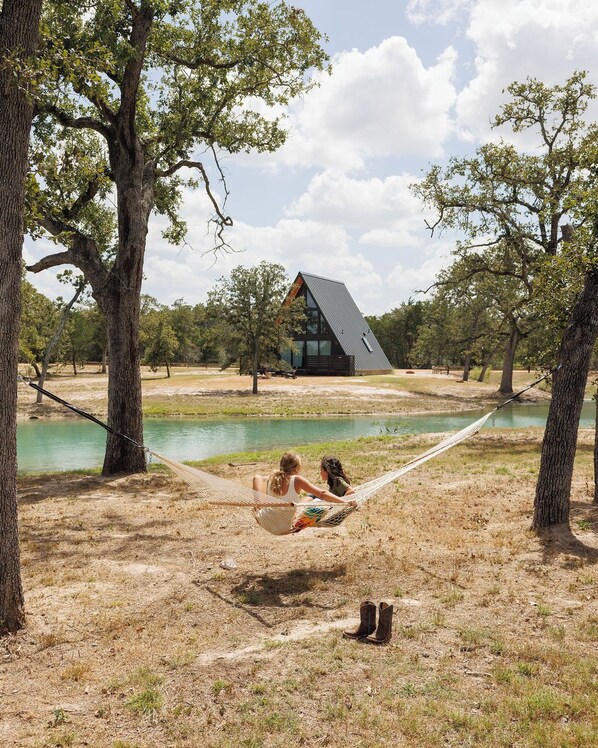 A hammock between trees with a lake and a modern house in the background.
