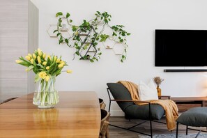Ein modernes Wohnzimmer mit einem Holzesstisch, einer Couch, einem an der Wand montierten Fernseher und Zimmerpflanzen.