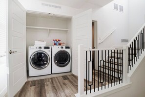 A laundry room with two front-loading washing machines, a shelf with laundry supplies, and a white staircase with a black railing. - Spacious & Stylish Rooftop Retreat l 2Bd/3Ba RiNo (Denver)