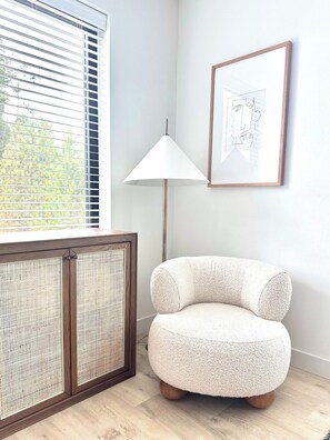 Een kamer met een raam, een staande lamp, een lijst met een afbeelding en een rond poef.