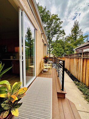 Une terrasse en bois avec une porte-fenêtre coulissante donnant sur une cuisine, le tout entouré d’une clôture en bois et de verdure.