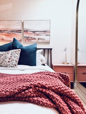 Une chambre à coucher avec un lit, des oreillers, une table de chevet, une lampe et deux tableaux accrochés au mur.