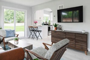A modern living room with a TV, seating, and a kitchen in the background. - Cottage Escape 1 BD w/Large Yard: Sloans Lake (Denver)
