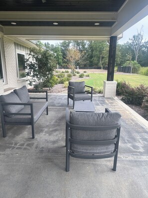 An outdoor patio area with a concrete floor, two grey upholstered chairs, a small table, and a sofa.