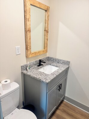 A bathroom with a granite countertop sink, a wooden-framed mirror, a toilet, and a trash can.