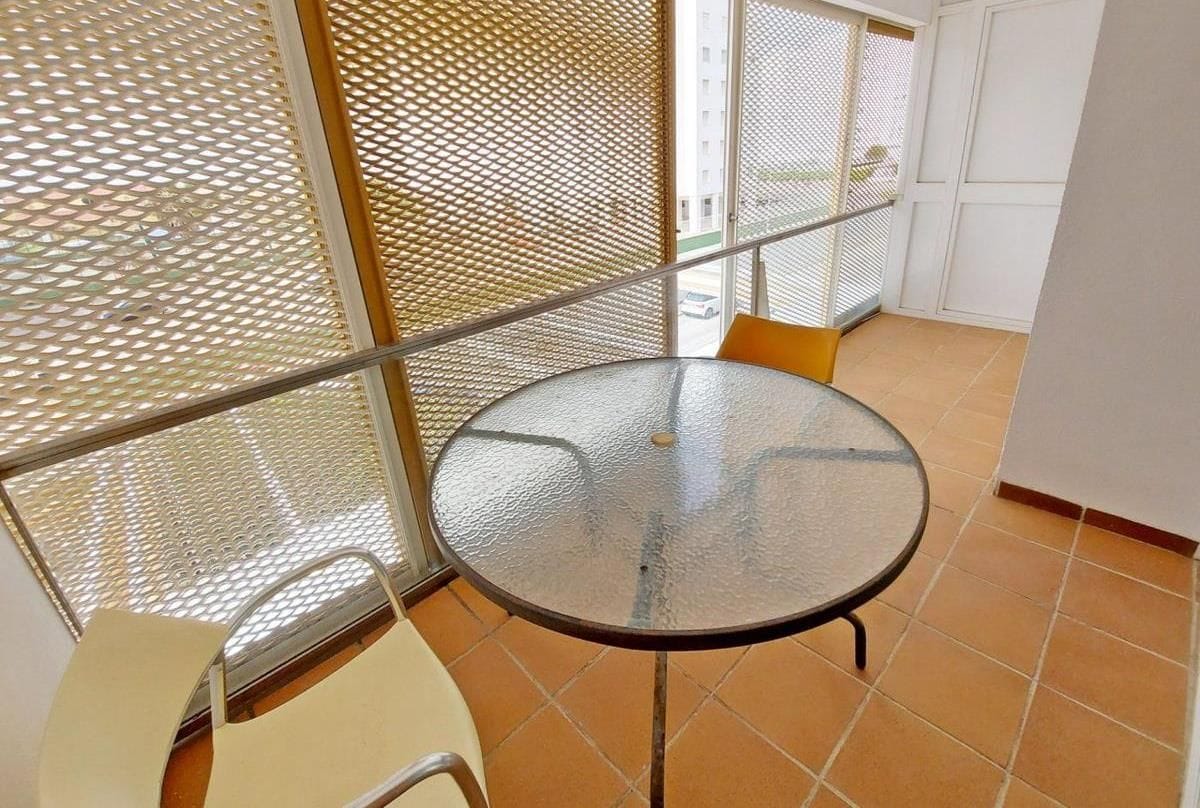 Una pequeña mesa redonda de cristal con patas metálicas, una silla blanca y un cojín amarillo, ubicada en una esquina con un separador de malla metálica y una puerta blanca al fondo.