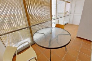 A small, round glass table with metal legs, a white chair, and a yellow cushion, situated in a corner with a metal mesh partition and a white door in the background.