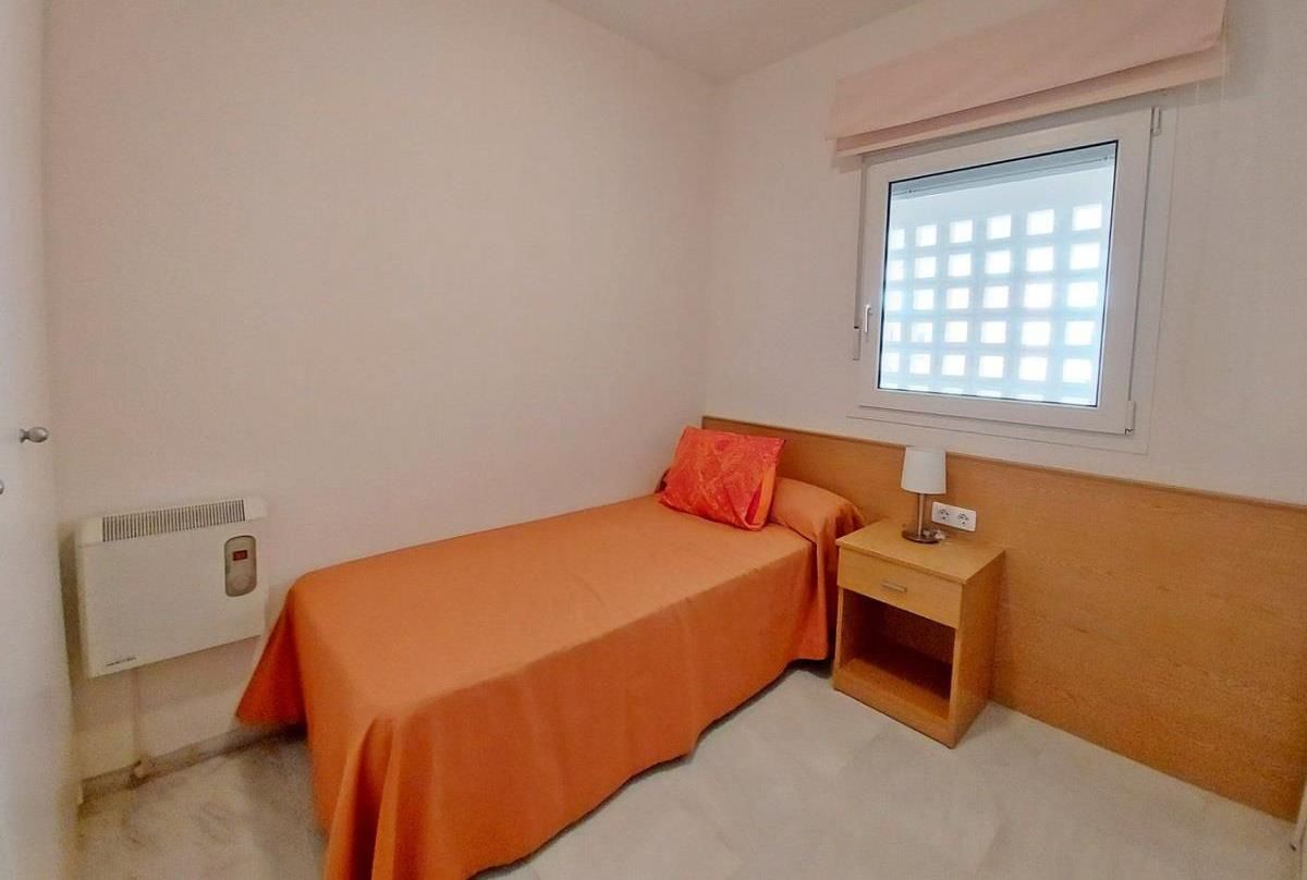 Una cama individual con una colcha naranja, una mesita de noche con una lámpara, una ventana con diseño de cuadrícula y un calentador fijado a la pared.