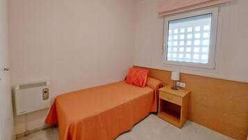 Una cama individual con una colcha naranja, una mesita de noche con una lámpara, una ventana con diseño de cuadrícula y un calentador fijado a la pared.