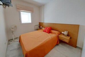 A bedroom with a bed, a bedside table, a lamp, a fan, and a window with a curtain.