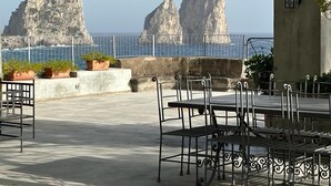 An outdoor seating area with tables and chairs, overlooking the sea with rocky formations in the distance. - TORRE SARACENA. Villa in Capri (M. Piccola) with beach access and a large terrace (Campania)