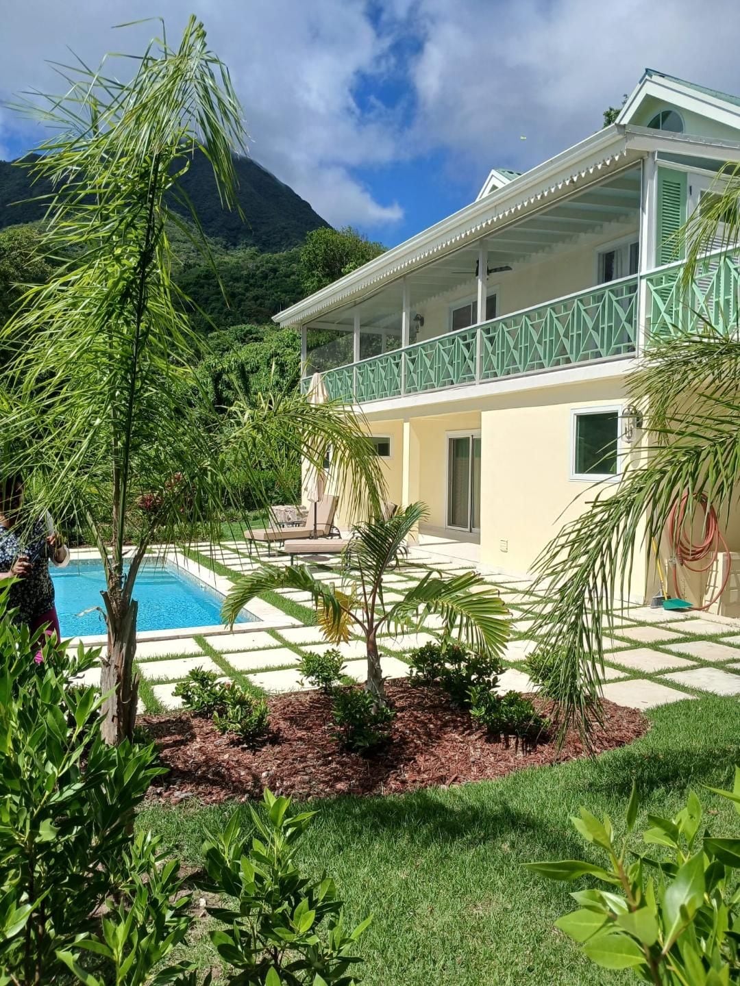 A two-story house with a pool, surrounded by lush greenery and a mountain in the background.