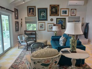 A living room with a sofa, armchair, and a table with a lamp. There are framed pictures on the wall and a window with a view. - Cozy 1-bedroom, 2 bath house  house in serene setting with AC (Pond Hill)