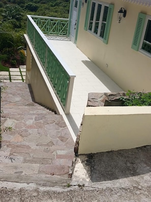 A stone staircase leading to a balcony with a green railing, a beige wall with shutters, and a stone planter with greenery. - Cozy 1-bedroom, 2 bath house  house in serene setting with AC (Pond Hill)