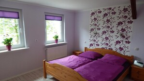 Et soveværelse med en træseng, to vinduer med skodder, en radiator, et natbord og en væg med blomstertapet.