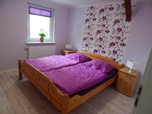 Une chambre avec un lit en bois, une fenêtre avec des stores, un radiateur, une table de chevet, une lampe et un papier peint à motifs floraux.