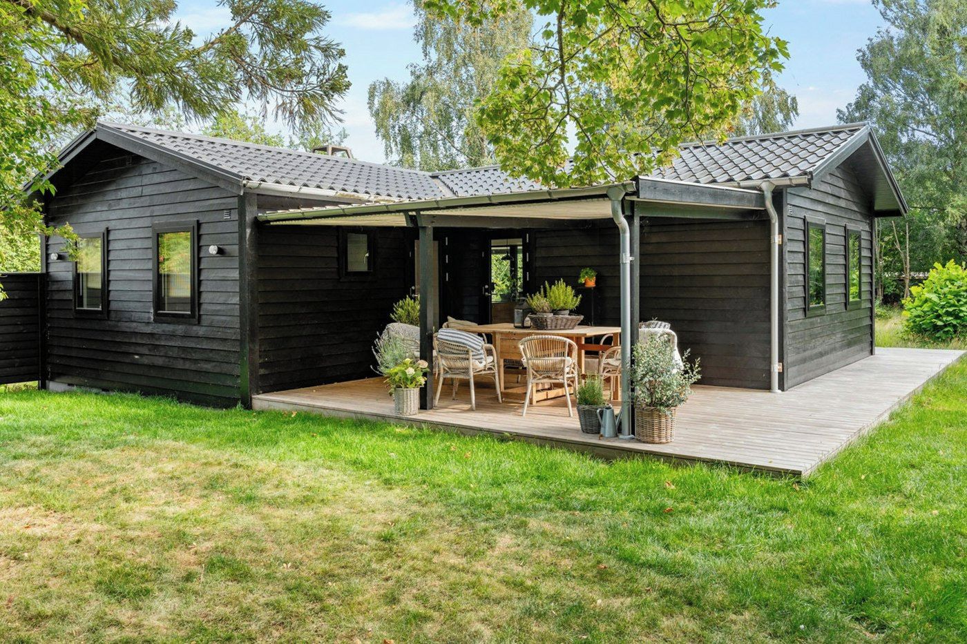 Een houten huis met twee verdiepingen, een overdekte patio, een eettafel met stoelen en potplanten.