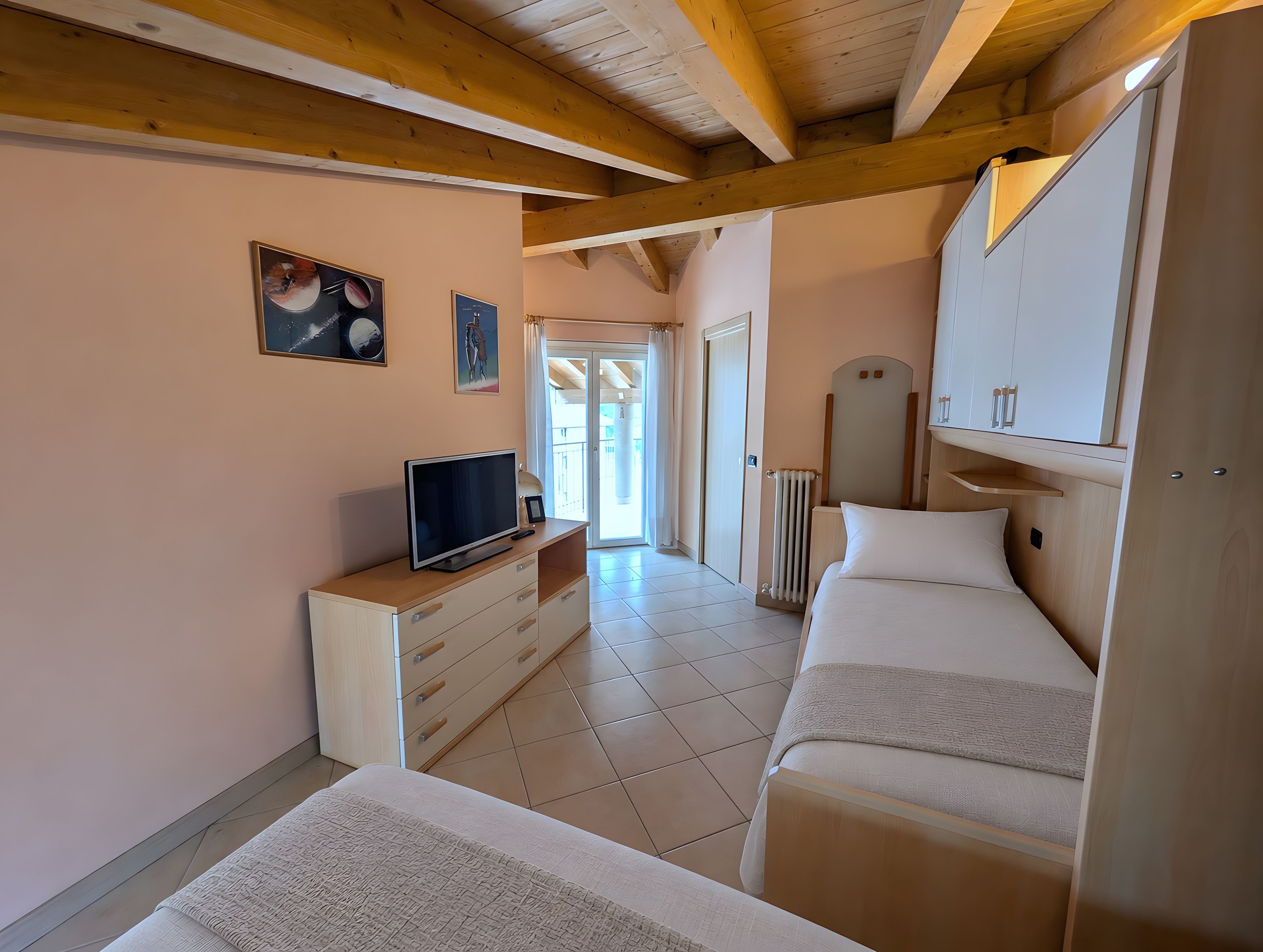 A hotel room with a bed, a TV on a wooden dresser, a radiator, and a door leading to a balcony.