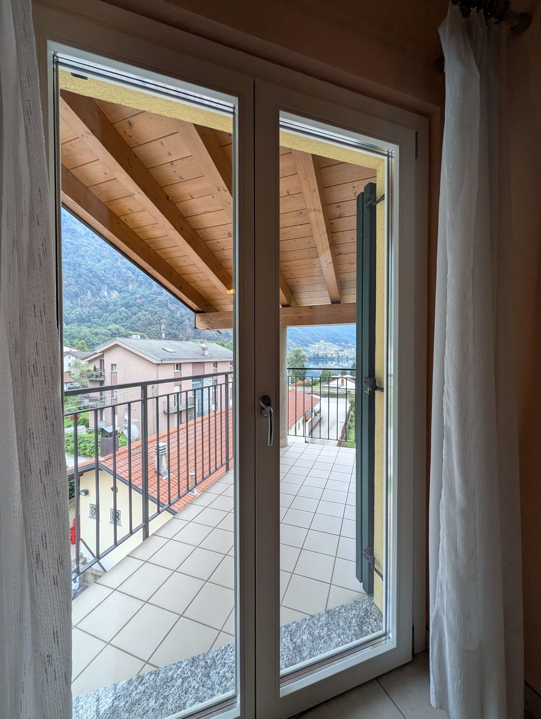 Una habitación con puerta corrediza de vidrio que da a un balcón con suelo de baldosas y vistas a casas y montañas.