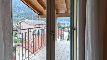 Una habitación con puerta corrediza de vidrio que da a un balcón con suelo de baldosas y vistas a casas y montañas.