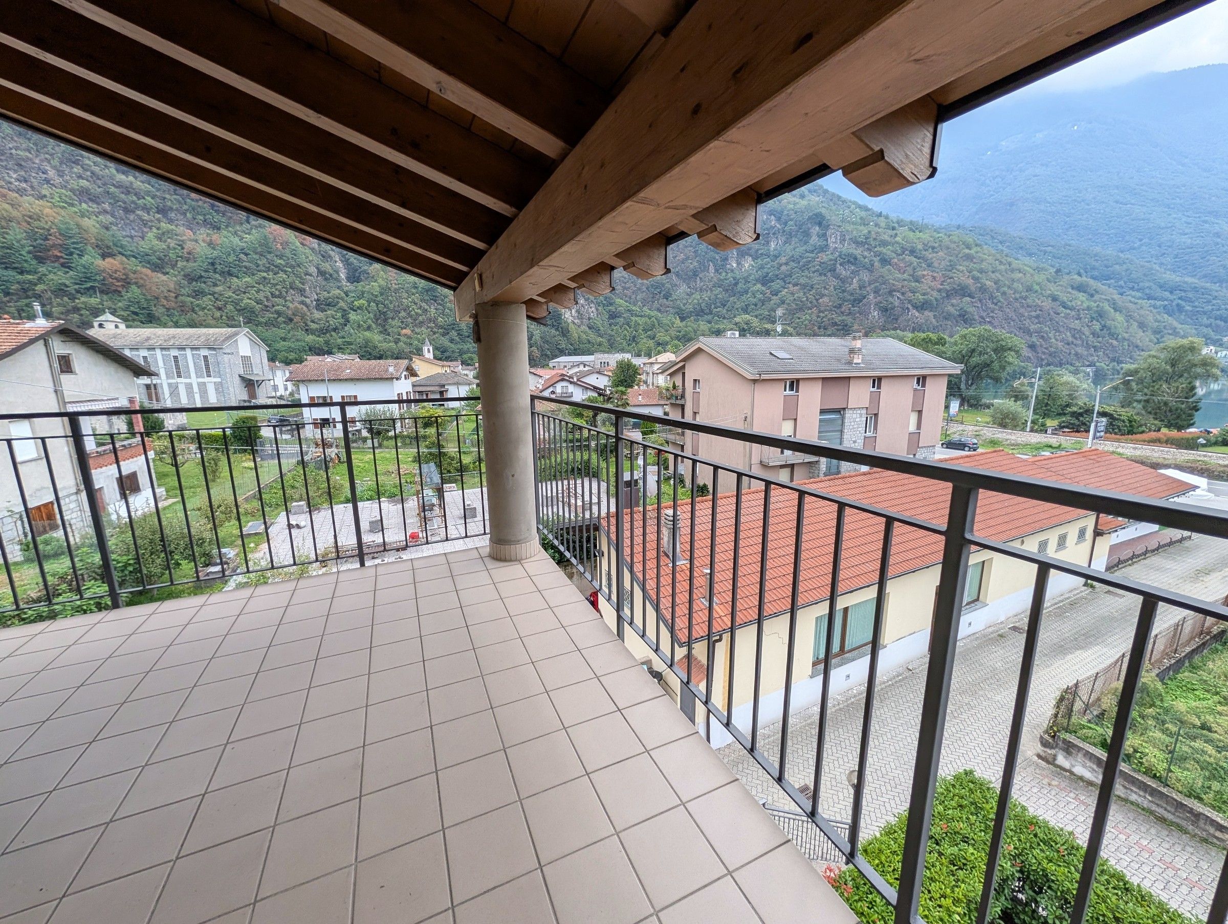 Un balcón con suelo de baldosas, techo de madera y vista a una zona residencial con casas y una montaña al fondo.