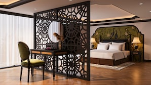 Habitación de hotel con cama, escritorio y silla, separador decorativo y un amplio ventanal.