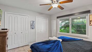 A bedroom with a bed, a ceiling fan, a window with a view of greenery, and white doors.