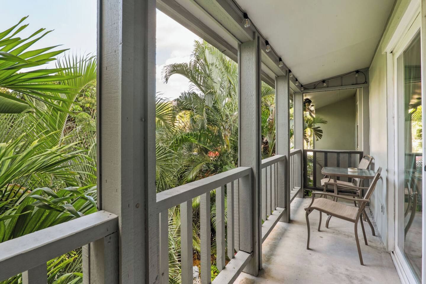 A covered balcony with a view of tropical plants, a table with chairs, and a glass door leading to the interior.