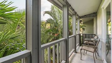 A covered balcony with a view of tropical plants, a table with chairs, and a glass door leading to the interior.