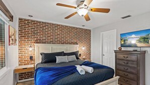 Un dormitorio con pared de ladrillo, ventilador de techo, cama con manta azul, cómoda y televisor en la pared.
