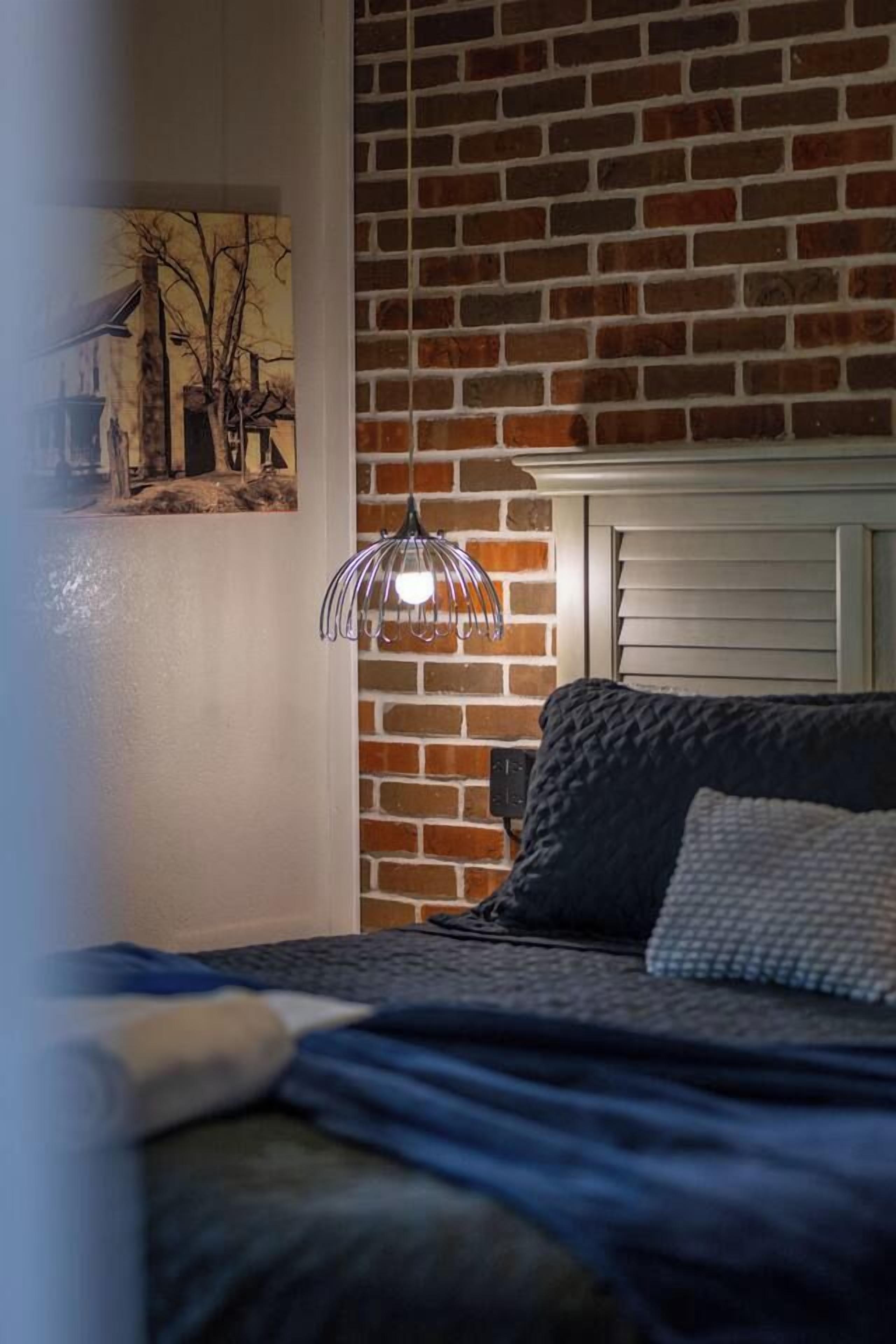 A bedroom with a brick wall, a bed with a dark cover, a hanging light fixture, and a painting on the wall.